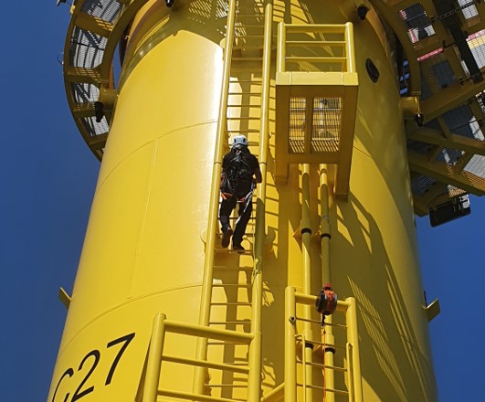 A wind turbine technician climbing the yellow foundation of an offshore wind turbine