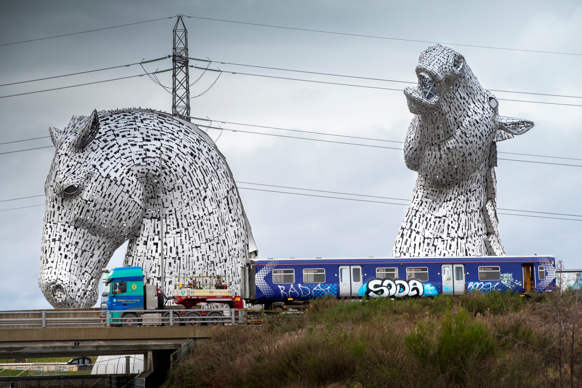 Image of Scotrail carraige loaded onto a truck, on its way to Bo-Ness, travelling past The Kelpies at Grangemouth, Forth & Clyde Canal.