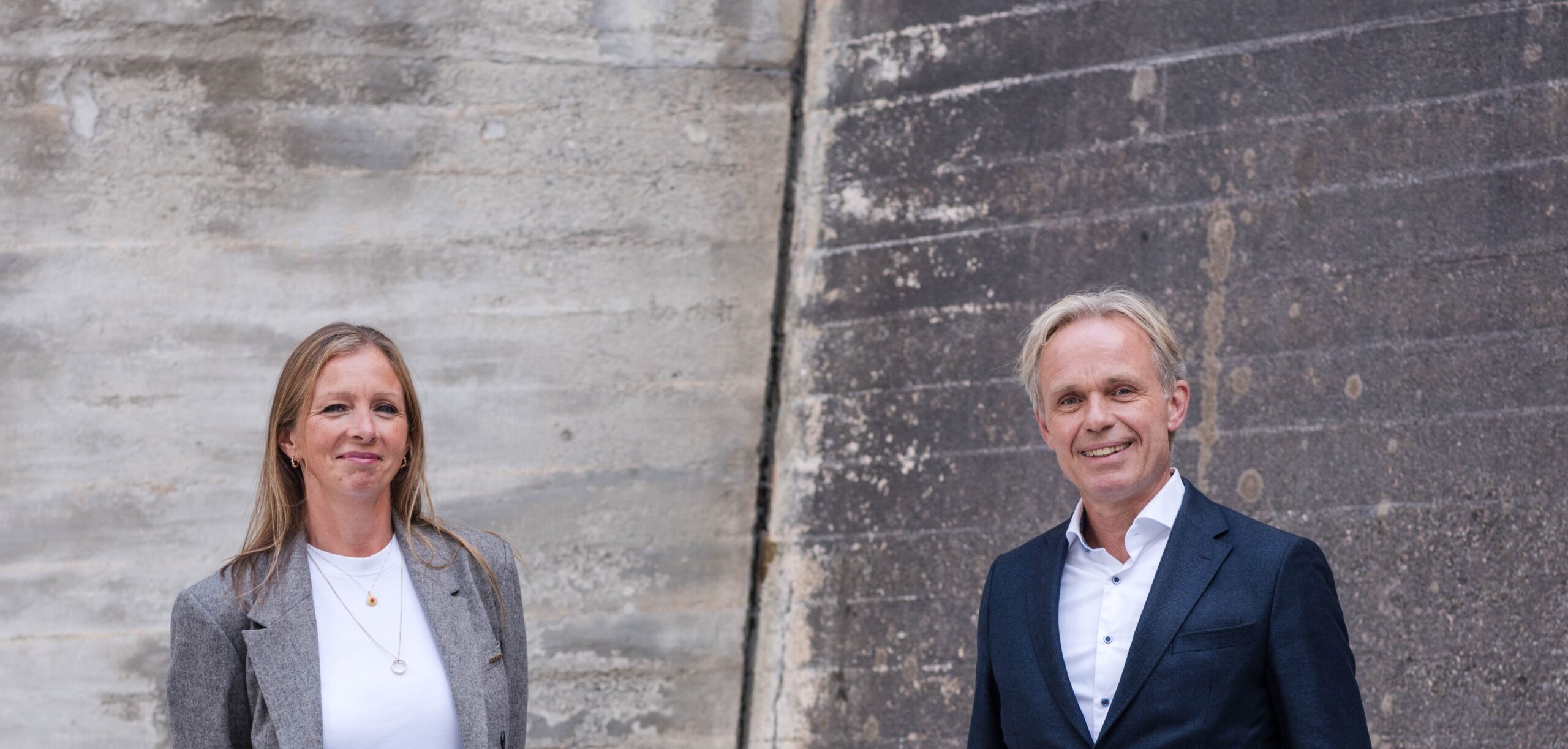 a woman and a man in suits standing next to each other in front of a concrete wall