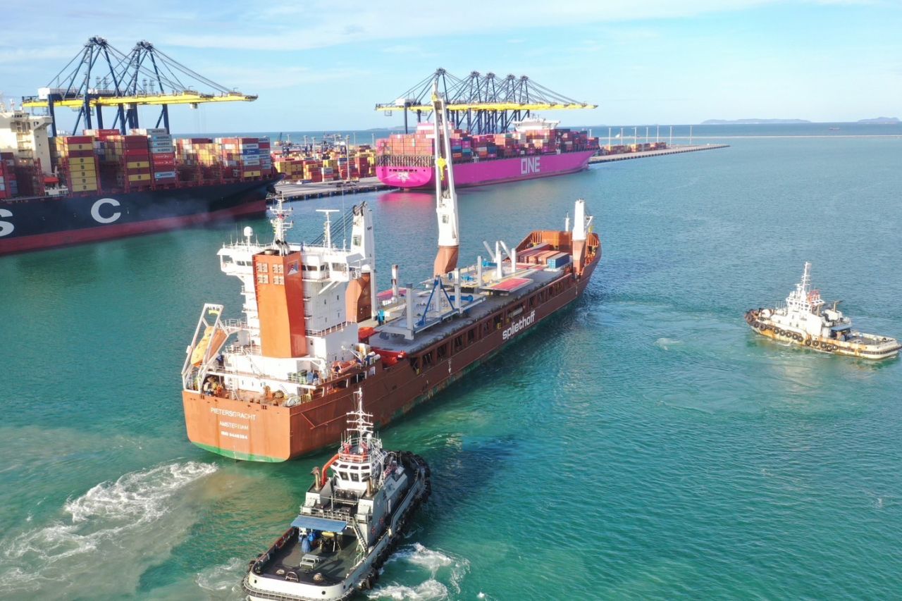 Large vessel surrounded by two tugboats with two other large vessels docked in the background