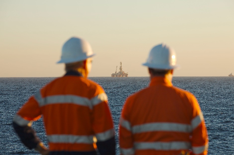 Two men in hard hats and jackets looking at an offshore platform in the distance