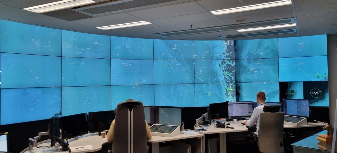 A large screen in a control room with two people sitting in front of it