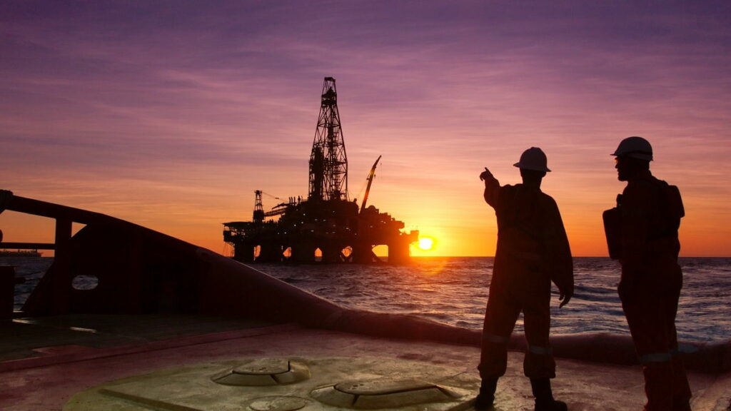 Two men standing on the coast at sunset with one of them pointing at an offshore rig