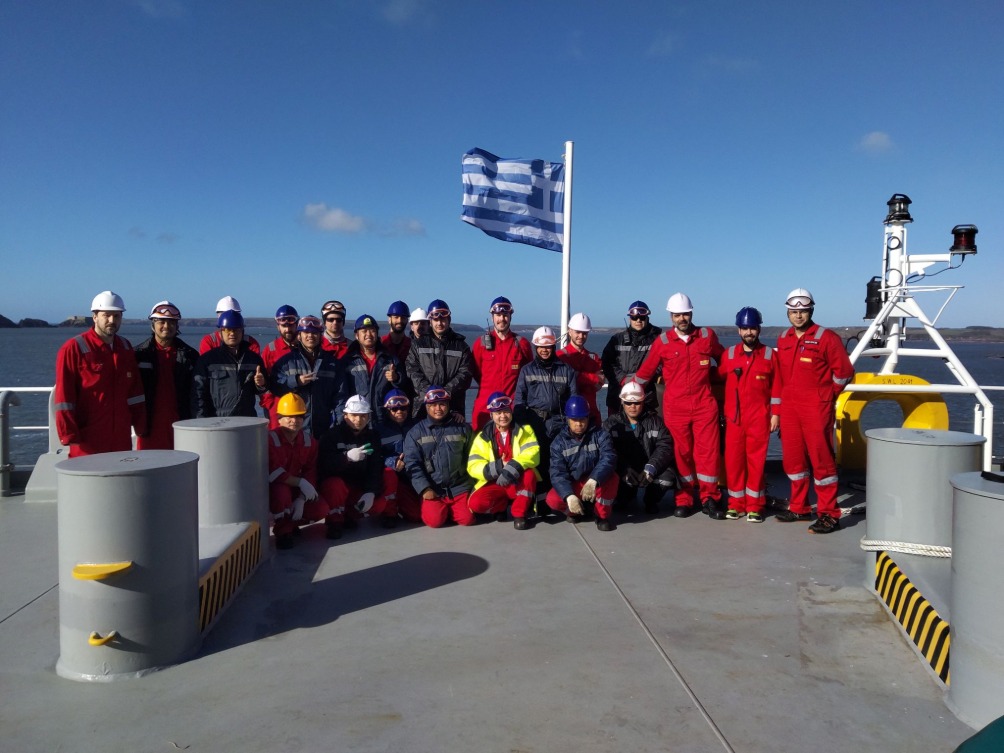 Several people standing in line on a vessel with Greek flag flying above them